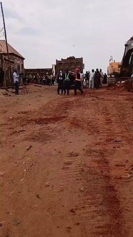 The video shows a construction site with an excavator and a dump truck in the background. There is a group of people gathered near the machinery, including some wearing construction gear and others in business attire. A man is standing with a camera and tripod. The ground is dirt and appears to be under construction. The sky is overcast. Three people are walking towards the camera. They include one man in a suit, one man in a black uniform, and a man wearing a safety vest and hardhat.