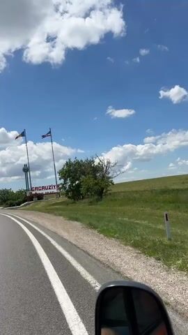 The video shows a view from a vehicle driving on a road. On the right side of the road, there is a large sign that says "Gagauziya" in red letters. There are two flags on tall poles next to the sign. The left flag is blue, yellow, and red, and the right flag is blue, red, and white. Behind the sign, there is a grassy hill with some trees. The sky is blue with white clouds. The road has a white dividing line. The video captures a brief moment of travel through this area.