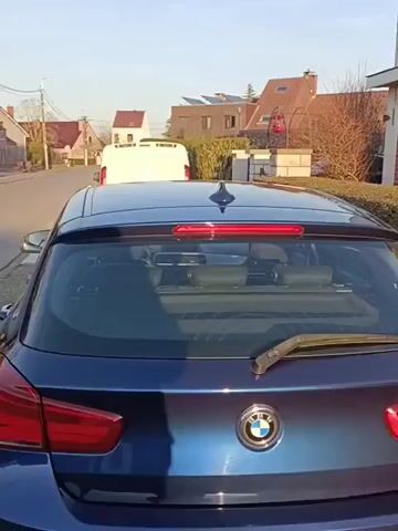 The video shows the back of a blue BMW 1 Series car parked on the side of a street. The car is facing away from the camera, and the camera slowly pans from the rear of the car down its side. The car is parked on gravel beside a sidewalk. Other cars and houses are visible in the background. The lighting suggests it is daytime. The car has a Belgian license plate.