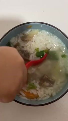 A hand holding a red spoon stirs a bowl of white rice soup. The soup contains pieces of meat, green vegetables, and sliced carrots. The bowl is blue with a dark rim. The background is a plain white surface.