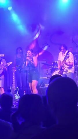 A live band performs on stage under blue stage lights. The lead singer, a woman with blonde hair, is in the center, wearing a black sequined top and black shorts, holding a microphone and singing. To her left, a man in a light-colored shirt and shorts plays a guitar. Behind her, another woman stands with a guitar, and further back, a drummer is visible behind a drum kit. To the right of the singer, a man in a light-colored shirt and shorts plays a guitar. In the foreground, the backs of audience members are visible, mostly out of focus. The stage has a backdrop with abstract designs. The lighting creates a smoky atmosphere.