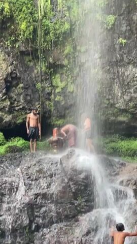 Here is a description of the video:

The video shows a small waterfall cascading over rocky terrain. Several people are present, including a shirtless man standing to the left of the falls, two people clustered near the top of the falls, and a young boy climbing on the rocks to the right. The surroundings include lush greenery and a rocky cliff face covered in moss.