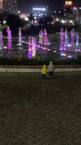 At night, two children stand on a paved area facing a fountain. The child on the left is wearing a bright yellow hooded jacket, while the child on the right is wearing a jacket with a light-colored pattern. They are both looking at the fountain, which has multiple jets of water illuminated with various colors. The jets of water shoot upwards in vertical streams. The paved area is decorated with a dark geometric pattern. Behind the fountain, there's a low wall with plants growing behind it. In the background, buildings are visible, including a hotel sign with the word "HOTEL" on it. Streetlights and other city lights are also visible in the background. The overall scene is colorful due to the lights on the fountain. The two children appear to be enjoying the sight of the fountain.
