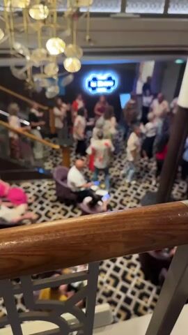The video shows a group of people dancing and socializing in an indoor space. The floor has a distinctive black and white leopard-like pattern. A neon blue sign reading "Le Bistro" is visible in the background. People are dressed in a variety of casual attire, with some wearing brightly colored, patterned shirts. A large, ornate chandelier hangs from the ceiling. Some individuals are seated in plush chairs, while others are standing and dancing. The atmosphere appears lively and celebratory. The camera is positioned from a higher vantage point, looking down into the scene, and the foreground is dominated by a wooden railing.
