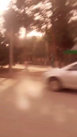 The video shows a street scene from a moving vehicle. There are trees lining the road, a building with a sign, and parked cars. The footage is somewhat blurry and has a warm tint.  The camera moves quickly.
