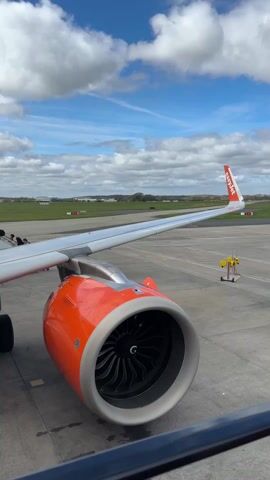Here's a description of the video:

The video shows a view of an EasyJet airplane engine and wing while passengers board. The engine is orange and gray, with visible fan blades. The wing is silver, and the EasyJet logo is visible on the tail fin. Passengers are walking up a set of stairs attached to the plane. In the background, there is a grassy airfield, a control tower, and a partly cloudy blue sky. The camera is positioned inside the aircraft, looking out at the scene. A black railing is visible at the bottom of the frame.