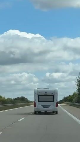 The video shows a white caravan being towed down a highway. The sky is blue with fluffy white clouds. The road is paved and has white lane markings. Trees and greenery are visible on the sides of the road. The camera appears to be positioned inside another vehicle, following the caravan. The caravan is the central focus of the shot.