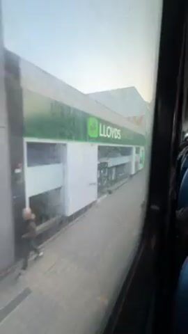From the window of a bus, a row of buildings along a street are visible. First is a Lloyds Pharmacy, followed by a shop with signage in Arabic script. Next is a pink-fronted "Cake Inn" with a closed shutter, and beyond that, a shop with an orange front, also shuttered.  The buildings are mostly white with a gray stone sidewalk. The sky is visible above, suggesting an early morning or late afternoon setting. Some people are visible sitting inside the bus.