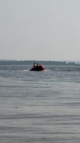 A small red inflatable boat with three people on board moves across a body of water. The boat is being operated by a person in a gray shirt and a hat. Two other people, appearing to be children, are seated in front of the operator. The water is calm with small ripples, and a distant tree line is visible on the horizon under a hazy sky. The boat is creating a wake as it moves.