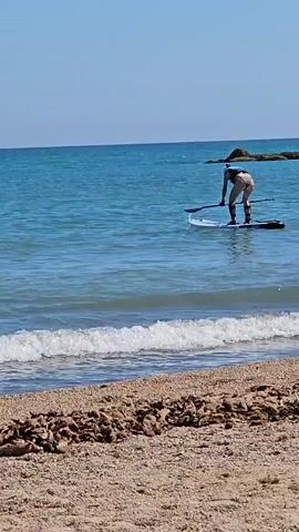 The video shows a person stand-up paddleboarding in the ocean on a sunny day. The person is wearing a life vest and is paddling with a single-bladed paddle. The water is blue and calm with small waves breaking on the shore. The beach in the foreground is sandy with some debris. The sky is clear and blue.