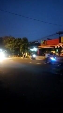 A handheld vertical video records a street scene at dusk. The sky is dark blue, transitioning to black. On the left side of the street, a vehicle with bright headlights moves towards the camera, its lights blurring the surrounding trees and foliage. Further down the street, the lights of buildings and street lamps illuminate the scene. A few structures are visible, including a building with a red awning and decorative banners. To the right of the street, another vehicle, possibly a car or motorcycle, with blue lights, drives away from the camera. The road surface is dark asphalt.