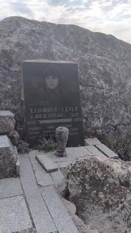 The video shows a tombstone with a portrait of a woman, her name, and dates of birth and death. The tombstone is set against a large rock formation. Steps made of stone lead up to the tombstone. The environment is rocky and appears to be outdoors with a cloudy sky.
