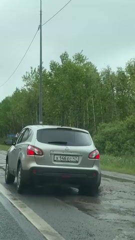 The video shows a silver Nissan Qashqai driving on a road with wet asphalt, with its rear facing the camera. The car has the license plate X 146 EC 47. The shoulder of the road is muddy, and the car splashes through a puddle as it pulls away. The surrounding environment includes green trees, a gray sky, and roadside elements like utility poles and signs.