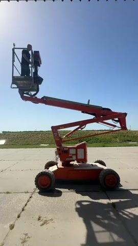 The video shows a person standing in the basket of an orange boom lift, which is elevated on a concrete surface. The boom lift has four large, knobby tires and an intricate arm structure that supports the basket. The person appears to be working on something within the basket. The background includes a strip of green vegetation and a clear blue sky with a bright sun. A partial shadow of an object offscreen is visible above the boom lift. The camera remains stationary throughout the video.