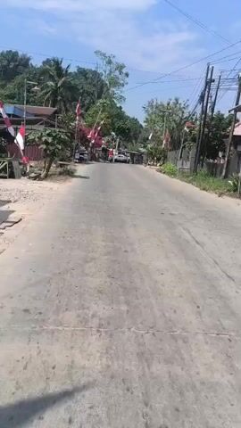 The video shows a paved road in a rural or suburban area. The road is lined with trees, some of which are bearing red flowers. Several Indonesian flags are displayed along the sides of the road, indicating a celebration or national event. There are a few buildings visible, and some parked motorcycles. In the distance, a white car is driving away from the camera. The sky is blue with some clouds. The overall impression is a sunny day in a community decorated for a national holiday.