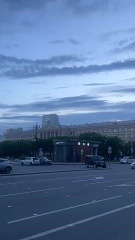 A wide shot captures an urban street scene in Khabarovsk, Russia, under a dramatic, cloudy sky at dusk. The sky is filled with layers of dark, elongated clouds, hinting at unsettled weather.

In the foreground, a multi-lane asphalt road takes up the lower half of the frame, with white lane markings visible. Several cars are on the road, including a white sedan in the right lane, a dark SUV in the middle lane, and a blue hatchback in the left lane, its brake lights illuminated. A white police car with the number "827" on its side is also present in the traffic.

Across the street, a modern, dark-colored kiosk or small building with glass windows stands prominently, adorned with a few illuminated star-like symbols. Behind this kiosk, a row of lush green trees lines the edge of a wide sidewalk. Beyond the trees, a grand, classical-style building with multiple stories, arches, and columns dominates the background, its architecture suggesting an important institution like a university or government building. Another taller, lighter-colored building rises above the main structure. A streetlamp is visible on the left side of the sidewalk.

The overall mood is subdued and slightly moody, typical of an overcast evening. The presence of the police car and the general urban setting suggest a normal city environment. The time of day is clearly evening, as indicated by the dim lighting and the dark clouds, with the city lights not yet fully illuminated. No people are distinctly visible in the frame, adding to the focus on the urban landscape and the architecture.