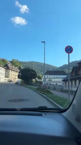 A view from inside a vehicle driving on a paved road in a European town. The buildings on the left are multi-story, with apartments or offices. The road curves slightly to the right, leading towards a wider street. On the right side of the road, there's a building with a modern architectural style, featuring large windows and a grey facade. A light pole stands tall next to it. Further down the street, more buildings and trees are visible, with a forested hill in the background under a clear blue sky with a single white cloud. The view through the windshield is unobstructed, allowing a clear perspective of the town. The side mirror on the right reflects the driver and part of the car's interior.