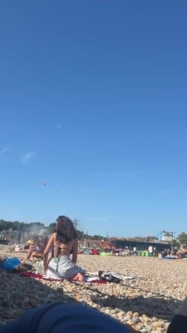 A group of people are on a pebble beach under a clear blue sky. In the foreground, a person's legs are visible, lying on the ground. In the mid-ground, a woman with dark, curly hair is sitting on a red blanket, facing away from the camera. She is wearing a bikini top and light-colored pants. To her left, another person is partially visible, also sitting on the blanket. Further back, other people are scattered on the beach. A kite is flying in the sky to the left of the frame, with a smaller kite or object attached to its string. In the background, buildings and trees can be seen. The sun is shining, casting shadows on the beach.