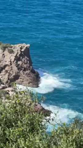 From an elevated position, the video shows a rugged, rocky cliff jutting into a vibrant blue ocean. Waves crash against the base of the cliff, creating white foam that contrasts with the deep blue of the water. In the foreground, green bushes and foliage partially obscure the view. The sky is a clear, bright blue, and the overall scene appears to be taken on a sunny day.