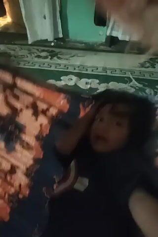 The video shows a child lying down, with their right hand touching their head. The child appears to be looking at the camera with a distressed expression. The child is wearing a dark t-shirt with a shield emblem on it. The child is lying on a patterned surface, possibly a bed or sofa, with a colorful blanket draped over their left side. In the background, there are white curtains and a wall painted green. The lighting is dim, suggesting it might be nighttime or indoors with the lights off.