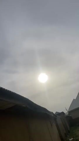 The video opens with a view of a bright, diffused sun high in a cloudy, grey sky. A dark, corrugated roofline is visible in the bottom left corner, suggesting a building below the camera's perspective. Faint outlines of distant structures can be seen on the horizon to the right. The sun's light creates a strong glare, indicating daytime. An audible sound of a vehicle engine, possibly a car or bus, and a horn is present throughout the initial frames. At the 0:03 mark, the camera quickly pans down or is covered, revealing a close-up of a plain, textured brown wall for the remainder of the video, while the engine sound persists. The location context is Tilde, Nigeria.