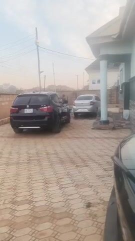 The scene is a driveway paved with hexagonal tiles, containing a black BMW SUV and a silver sedan parked near a house. The house has green trim and white pillars supporting the roof of the porch. A person walks between the two cars. Power lines stretch across the sky, which is hazy. A portion of another black vehicle is visible in the foreground.