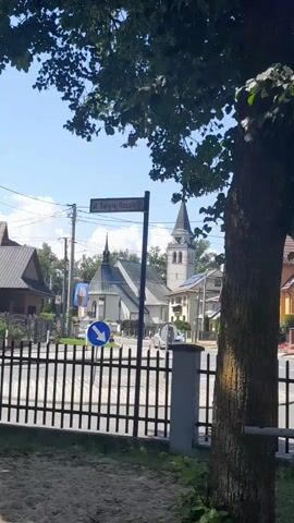 The video shows a street scene with a fence in the foreground. A street sign reads "ul. Swietej Rozarii." Beyond the fence, there is a building with a steeple, likely a church. Another building with a steeple is visible to the right, and residential buildings are on either side of the street. A blue and white directional sign is visible on a pole. Cars are present on the road, and trees are in the foreground and background. The sky is blue with some clouds.
