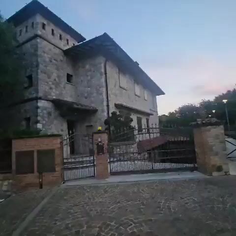 The video pans across the front of a large stone building with a tower on the left. The building has multiple windows and a pitched roof. In front of the building is a metal gate, a stone wall, and a paved driveway. To the right of the building, there is another gate, a brick wall, and trees. The sky is visible and appears to be dusk.