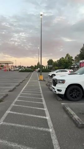 The video shows a parking lot at dusk. The sky is partly cloudy with soft pink and orange hues from the setting sun. A tall lamppost stands in the middle of the frame, and another is visible in the distance. Several cars are parked in the lot, including a white pickup truck in the foreground on the right. The truck has black rims and tinted windows. To the left of the pickup truck, a white SUV is parked. Further in the background, there is a gas station with red and white signage, and trees line the perimeter of the lot. The parking spaces are clearly marked with white lines, and concrete parking blocks are visible in front of the spaces. The overall atmosphere is calm and quiet.