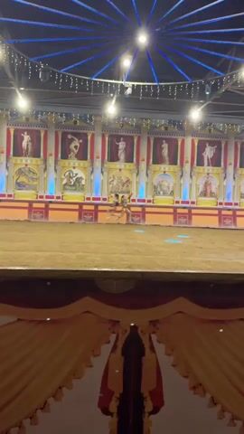 The video shows a performance in an arena. The arena floor is covered in sand. The backdrop is a painted mural depicting Roman-style architecture and scenes, with statues and columns. There are two women on the sand, dressed in what appears to be gladiator-like costumes. They are both wearing short, ornate skirts and upper body armor. The woman on the left has her hair in a ponytail and is wearing a red headband. The woman on the right has her hair down. Both women appear to be moving rapidly across the sand, possibly engaged in a choreographed fight or demonstration. Blue spotlights are projected onto the sand. The arena is covered by a ceiling with exposed metal beams and a circular light fixture. In the foreground, there is a decorative fabric draped across the bottom of the frame, resembling the front of a stage or seating area. The overall impression is of a historical re-enactment or performance.