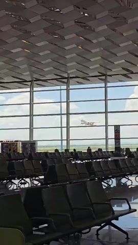 The video captures the interior of an airport terminal with rows of empty green seats in the foreground. Large windows reveal an outdoor scene where an airplane is ascending into a cloudy sky. A person wearing a dark skirt and a light-colored top, with a blue cap, walks across the frame from left to right, passing the empty seats. The ceiling of the terminal features a distinctive, geometric pattern.