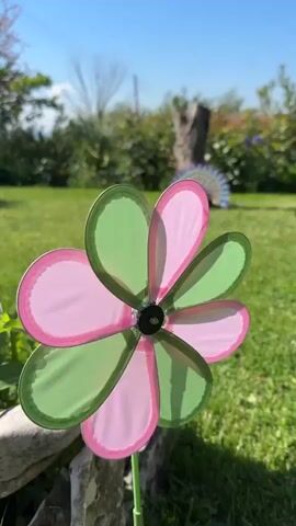 The video presents a close-up of a pinwheel-shaped garden ornament, with alternating pink and green plastic petals, situated in a grassy garden on a sunny day. The pinwheel is still, not spinning. Behind it, in the blurred background, are other garden features like shrubs, a tree trunk, and a partial view of a peacock’s tail. The pinwheel is positioned in front of a rocky edge, and some leaves are visible near its base. The sky above is clear and blue.
