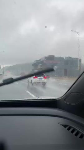 The media file shows a view from inside a car driving on a highway during a heavy rainstorm. The windshield is wet, with water droplets and streaks obscuring the view. A windshield wiper is partially visible on the left side of the frame, in motion.

The main subjects are vehicles on the highway. A white SUV is prominently featured in front of the car, its taillights visible. Further ahead, a red bus and other vehicles are seen through the rain. The scene is a multi-lane highway with a concrete barrier separating lanes. In the background, a large, modern building complex is visible, with some signage.

The activity is driving on a highway under adverse weather conditions. The weather is clearly rainy and overcast, creating a gloomy atmosphere. The time of day appears to be daytime, given the visibility, but the heavy clouds suggest it might be late afternoon or early morning.

Notable details include the water splashing from the tires of the vehicles on the wet road, indicating heavy rain and poor visibility. The building in the background has some visible text, but it is blurry and difficult to read due to the rain and distance. The overall mood conveyed is one of caution and perhaps a bit of frustration due to the challenging driving conditions. The location context is Yantian District, China, which is a coastal district in Shenzhen known for its port and industrial areas.