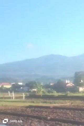 The video shows a blurry outdoor scene with a clear blue sky and a hazy mountain range in the background. In the foreground, there is a field with tilled soil. In the middle ground, there are several houses with red roofs, trees, and some electrical poles. A watermark in the bottom left corner reads "OPPO A76". The camera appears to be stationary. There are no visible people or animals. The overall lighting suggests it is daytime, possibly morning or late afternoon due to the haze.