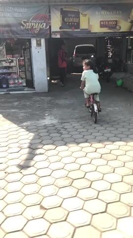 A young girl with dark hair, wearing a light green outfit, rides a pink bicycle towards the right of the frame. She is positioned on a patterned stone walkway. Behind her, an adult wearing a red shirt and dark pants walks away from the camera. A dark-colored van is parked in the background, partially obscured by a building with signs. The environment appears to be an outdoor commercial area with storefronts visible. Sunlight casts shadows on the walkway.