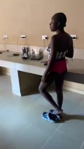 A person, likely female, with dark skin and short black hair, is seen from the back. They are wearing a black and white checked top and pink bottoms, and blue Crocs. They are standing in a room with cream-colored walls and light tile flooring. A long table runs along the wall, with multiple electrical outlets and cords plugged in. There are various items on the table, including phones and a laptop. A window with bars is visible in the background. The person walks across the room, moving the camera.