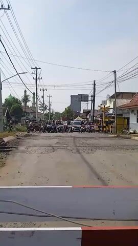 The video shows a street scene at a railroad crossing in what appears to be a small town. The crossing gate is down, blocking traffic, and a warning alarm is sounding.  A group of motorcycles and a few cars are stopped, waiting for the train. The area around the crossing is somewhat dusty and under construction. Overhead, there are utility poles and wires.  On the right side of the frame is a small building with a red tile roof, likely associated with the railroad. In the distance, a mosque is visible to the left, with other buildings making up the townscape.  The sky is clear and bright.