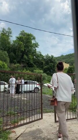 Two women walk towards an open, rusty metal gate. The woman in front is wearing a white shirt and beige pants, carrying a brown purse. The woman behind her also wears a white shirt and beige pants, with a black crossbody bag. Through the gate, several people are gathered in what appears to be a parking area. There are at least two white cars visible, and a person on a bicycle. The background is filled with lush green trees and vegetation on a hillside, suggesting a rural or semi-rural setting. The sky is cloudy. The overall impression is a casual gathering or arrival at a location, possibly a residence or event space. The time of day appears to be daytime, given the lighting.