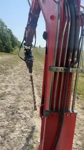 This media file shows a close-up view of a red excavator attachment, specifically a drill auger, in the ground. The auger is designed for digging holes, likely for planting posts or vines. In the background, a vineyard is visible, with rows of young grapevines supported by stakes. The scene suggests agricultural work in progress, possibly the establishment or maintenance of a vineyard.

The footage shifts to an interior view of a vehicle's side mirror. Through the mirror, the same vineyard landscape is reflected, providing a wider perspective of the rolling hills and lush greenery. The clear blue sky indicates a sunny day, likely in the late morning or afternoon, with good weather for outdoor work.

The setting appears to be a rural area with cultivated land. The presence of the vineyard and the type of machinery used point towards a farming or viticulture operation. The overall mood is one of productive activity, with the focus on the task at hand and the surrounding natural environment. No people are explicitly visible in the footage, but their presence is implied by the operation of the machinery. The location context provided is Gravesham, United Kingdom.