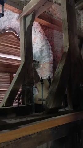 The video shows an interior shot of a bell tower. The camera is positioned at eye level, looking upwards at a large bronze bell suspended from heavy wooden beams. The bell is ornate and appears to be quite old. The wooden structure supporting the bell is made of thick, rough-hewn timbers, some of which are intricately carved. The background shows a brick wall with arched openings, suggesting the bell tower is part of a historical building. The lighting is dim, with some natural light filtering in from the openings. There are no people visible in the video, and no actions are taking place other than the slight movement of the camera. The overall impression is one of age, grandeur, and perhaps a sense of quiet anticipation.
