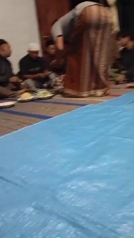 A group of men are sitting on the floor around a blue tarp. There are plates of food and cups on the tarp. One man is standing and wearing a brown and white striped sarong. The other men are wearing various traditional clothing, including skullcaps and sarongs. Some of the men are eating and talking. The environment appears to be indoors, possibly a home.