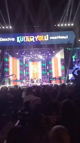 The video shows an outdoor concert stage at the Türkiye Kültür Yolu Festivali, viewed from the audience's perspective. The stage is elaborately lit with colorful lights and features a large screen that displays images of the performers.  A band is performing on stage, and their performance is projected on the screen. A large crowd of people is visible in front of the stage, many of whom are raising their hands. The event appears to be well-attended, with a lively atmosphere created by the music and light displays. Outside the venue, there is a street with passing cars.