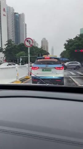 The video shows a view from inside a car looking forward at traffic on a wet city street under an overcast sky. In the immediate foreground, the dashboard and windshield wipers of the car are visible. Ahead, a blue and white taxi is stopped in traffic, with its hazard lights illuminated. Behind the taxi, another light blue car is turning. Further ahead, traffic lights are red, and the street is slick with rain. Tall modern buildings, including a very tall skyscraper with a distinctive pointed top, line the street, interspersed with green trees. A red traffic sign indicating "no left turn" is visible on a pole to the left of the taxi. The overall impression is a busy urban scene during a rainy day.