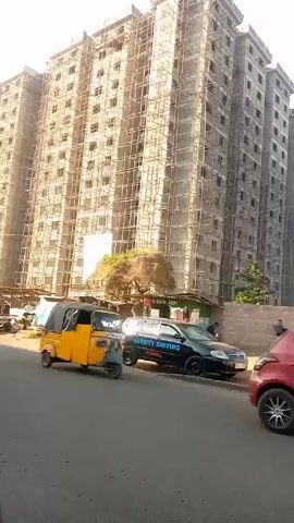 The video shows a street scene in front of a tall building under construction. The building is surrounded by scaffolding. Several people are visible walking on the sidewalk, and a yellow auto rickshaw is parked on the street. A black car with the words "VARSITY DRIVING SCHOOL" is also parked nearby. A silver SUV passes by the camera's vantage point.