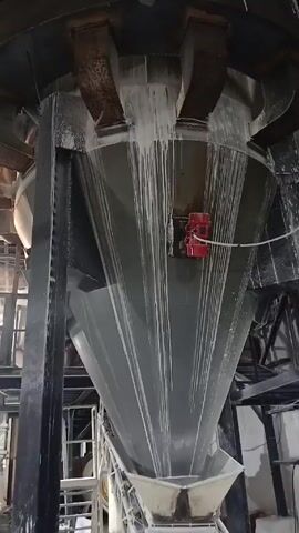 The video shows the interior of what appears to be a factory or industrial facility. The primary focus is on a large, conical hopper or funnel-shaped structure, covered with white residue. A conveyor belt runs below it, collecting material from the hopper. The structure is supported by black metal beams.

In the background, workers are visible in a wide-open room with multiple levels. They are moving around yellow bags, possibly filled with a material related to the production process. Some workers are wearing masks. The floor is covered in a layer of the same white residue as the hopper. Other industrial equipment is also visible in the background.