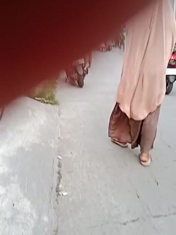 The video shows a street scene with several people and vehicles. In the foreground, a bicycle wheel is partially visible on the left. A person wearing a hijab stands near a scooter on the left side of the street. Further down the street, a person in a brown robe and hijab is walking away from the camera, appearing to be heavily pregnant or carrying something large. Behind them, a man in a green shirt and light-colored pants is standing near a scooter. The background depicts a street with buildings, market stalls, and utility poles. The ground is paved, and there is some grass growing along the edge of the sidewalk. The overall impression is a bustling, everyday street in a residential or market area.