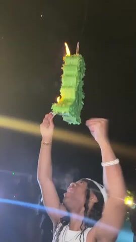 A man is holding up a green piñata shaped like the letter "L", which is on fire, with smoke rising from it. The man is wearing a white tank top, jewelry, and has long hair. He is looking up at the piñata with a smile, and has his arms raised, making fists. There are other people in the background watching, some of whom are smiling and laughing. There are also lights shining on the scene. The sky is dark.