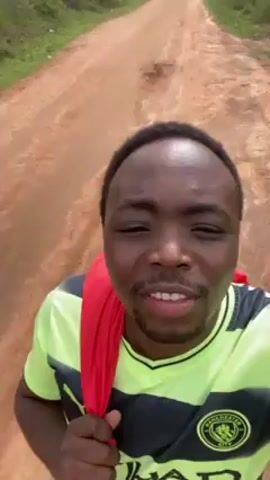 The video shows a man standing on a dirt road, talking to the camera. He is wearing a light green and black Manchester City soccer jersey and has a red cloth draped over his shoulder. He is smiling and appears to be in good spirits. The road is unpaved and stretches out in front of him, with vegetation on either side.