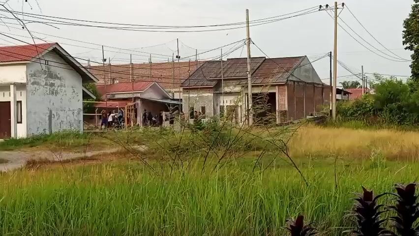 A group of people are gathered in front of a building. The building appears to be under construction or renovation, with scaffolding visible on the roof. There are several motorcycles parked nearby. The area is overgrown with tall grass and vegetation, and there are power lines running overhead. The sky is overcast.