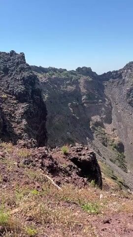 The video shows a view into the crater of a volcano. The crater is large and bowl-shaped, with steep, rocky walls. The bottom of the crater is covered in loose rocks and some sparse vegetation. The sky above is clear and blue. The camera appears to be positioned on the rim of the crater, looking down into it. The overall impression is one of a vast, desolate, and powerful natural landscape.